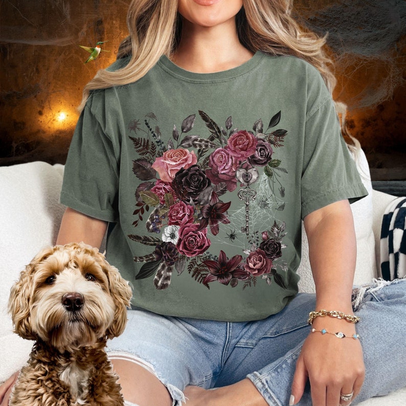 May include: A green t-shirt with a floral design featuring dark pink, red, and purple flowers, leaves, and feathers. A silver key and spider web are also included in the design.