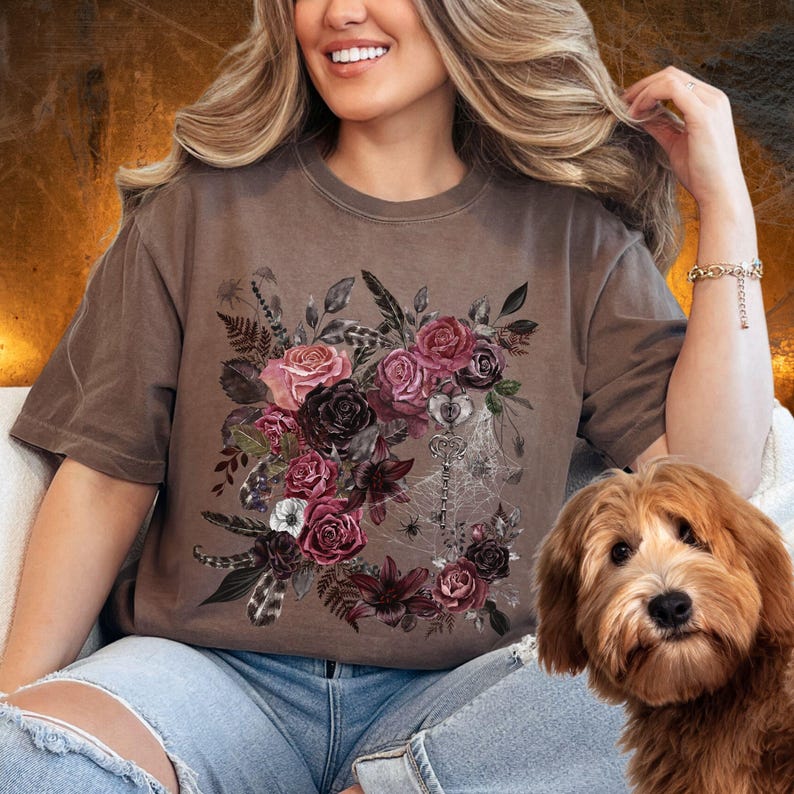 May include: A brown t-shirt with a floral design featuring dark pink, white, and black flowers, leaves, and feathers. The design also includes a key and spiderwebs.