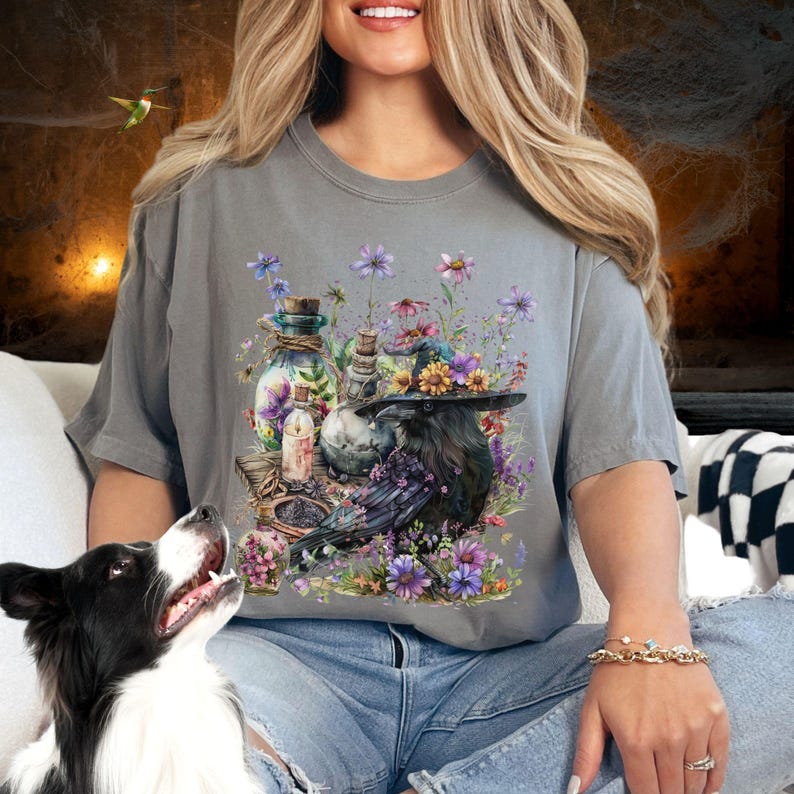 May include: A gray t-shirt with a watercolor design of a raven wearing a witch's hat surrounded by flowers and potion bottles.