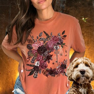 May include: A burnt orange t-shirt with a floral design featuring dark red, pink, and white roses, feathers, and a key with a spider web.