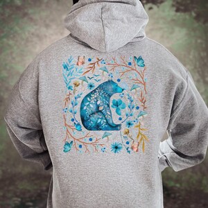May include: A gray hooded sweatshirt featuring a blue bear design. The bear is surrounded by a floral wreath with blue flowers, butterflies, and mushrooms. The design is centered on the back of the sweatshirt.