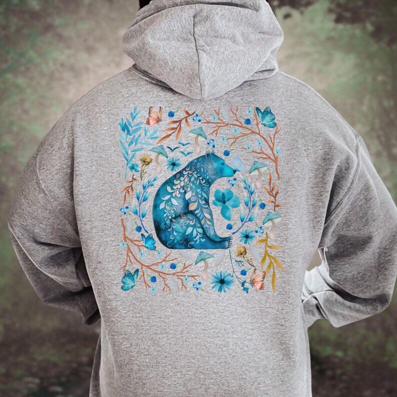 May include: A gray hooded sweatshirt featuring a blue bear design. The bear is surrounded by a floral wreath with blue flowers, butterflies, and mushrooms. The design is centered on the back of the sweatshirt.