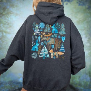 May include: Dark gray hoodie featuring a whimsical winter design on the back. The design includes blue rabbits, trees, a moose, and a fox. The artwork has a watercolor style with blue, white, and brown accents. The hoodie has a hood and long sleeves.