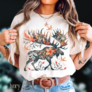 May include: Ivory-colored t-shirt featuring a detailed moose design with floral and leaf patterns in shades of orange, green, and brown. The moose is the central focus, with decorative elements around it. The word "Ivory" is printed at the bottom.
