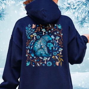 May include: Navy blue hoodie featuring a detailed illustration of a blue bear surrounded by flowers, mushrooms, and butterflies. The design is framed by branches and leaves in shades of brown and blue. The hoodie has a hood and long sleeves.