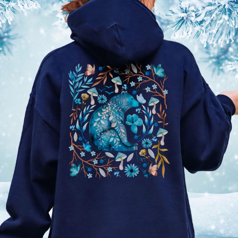 May include: Navy blue hoodie featuring a detailed illustration of a blue bear surrounded by flowers, mushrooms, and butterflies. The design is framed by branches and leaves in shades of brown and blue. The hoodie has a hood and long sleeves.