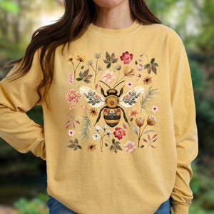 May include: A mustard yellow long-sleeve sweatshirt featuring a central graphic of a bee with intricate floral patterns on its wings and body, surrounded by a variety of botanical illustrations including small flowers, leaves, and stems.