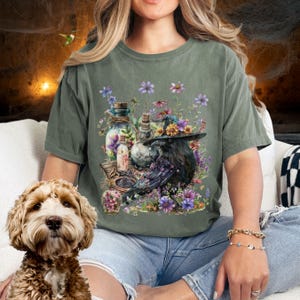 May include: A green t-shirt with a black raven wearing a floral crown and holding a potion bottle. The raven is surrounded by flowers and other potion bottles.