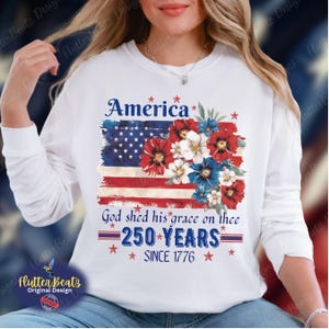 May include: White long-sleeve sweatshirt with a patriotic design. The graphic features an American flag, floral arrangement, and text that reads "America," "God shed his grace on thee," and "250 YEARS SINCE 1776."