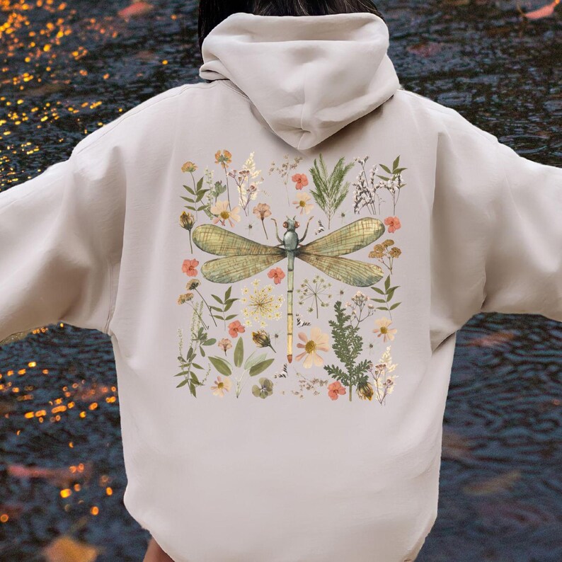 May include: Beige hoodie featuring a large dragonfly illustration with floral accents on the back. The dragonfly has green and gold wings, and the flowers are in shades of peach, yellow, and green. The hoodie has a drawstring hood and long sleeves.