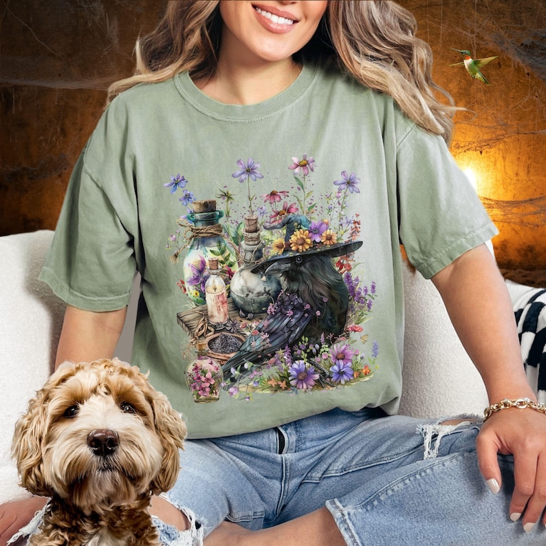 May include: A green t-shirt with a watercolor illustration of a raven wearing a flower crown and holding a potion bottle. The raven is surrounded by flowers and other potion bottles.