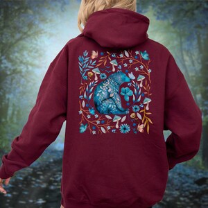 May include: A maroon hoodie featuring a detailed illustration on the back. The design showcases a blue bear surrounded by blue flowers, mushrooms, and butterflies. The hoodie has a hood and long sleeves, perfect for casual wear.