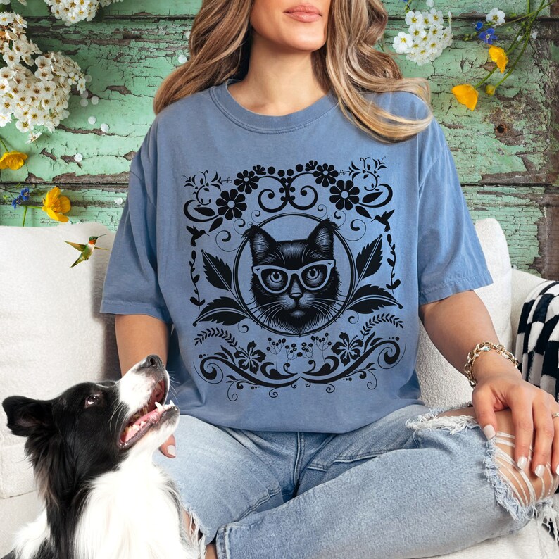 May include: A blue t-shirt with a black cat wearing glasses and a floral design. The cat is surrounded by a black floral pattern.