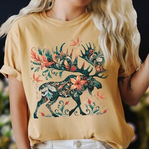 May include: Mustard yellow t-shirt featuring a detailed moose design. The moose is adorned with floral and leaf patterns in teal, orange, and green. Small birds and floral accents are scattered around the moose, creating a nature-inspired theme.