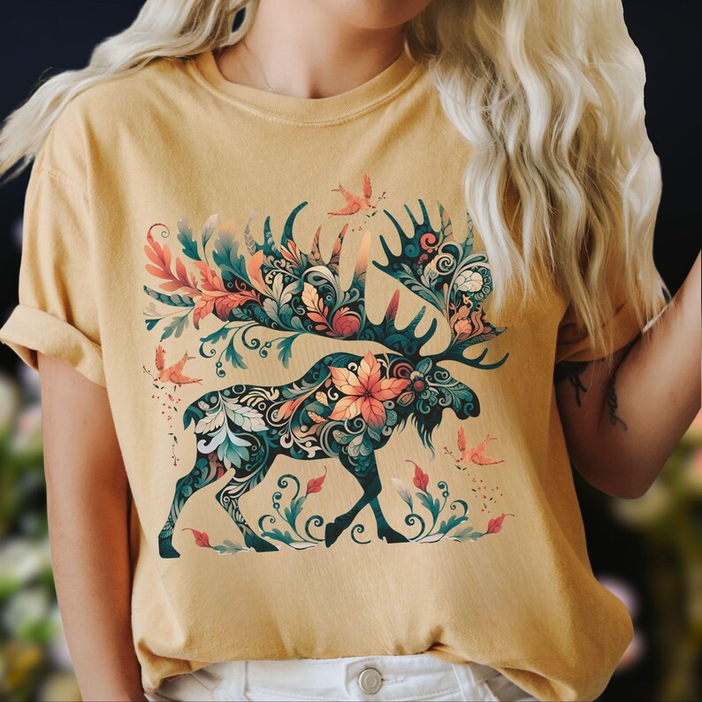 May include: Mustard yellow t-shirt featuring a detailed moose design. The moose is adorned with floral and leaf patterns in teal, orange, and green. Small birds and floral accents are scattered around the moose, creating a nature-inspired theme.
