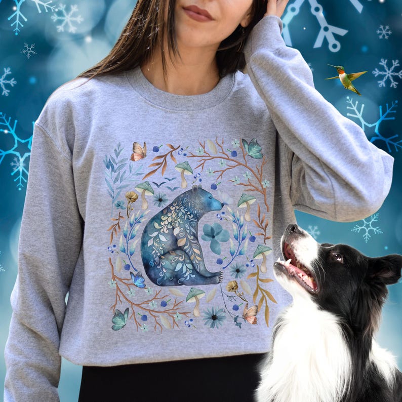 May include: A gray sweatshirt with a watercolor design of a bear sitting in a circle of flowers, leaves, and mushrooms. The bear is blue and gray with intricate details.
