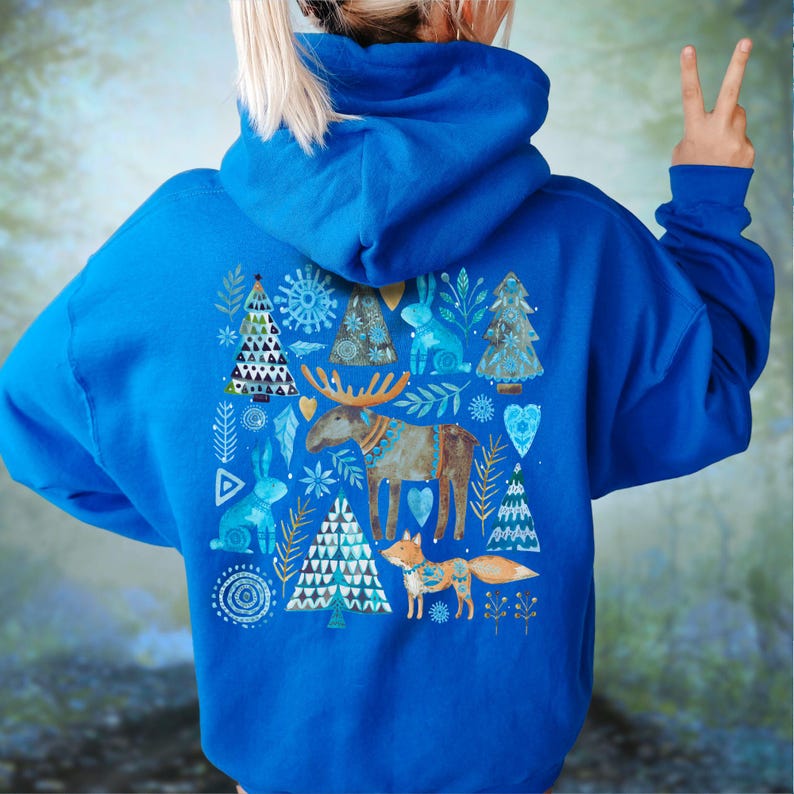 May include: Royal blue hoodie with a whimsical winter-themed design on the back. The design features a moose, fox, rabbits, and Christmas trees in shades of blue, gold, and white. The hoodie has a hood and long sleeves.