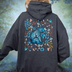 May include: Dark gray hoodie featuring a blue bear design surrounded by flowers, mushrooms, and butterflies. The bear is intricately detailed with floral patterns. The design is centered on the back of the hoodie, creating a nature-inspired aesthetic.