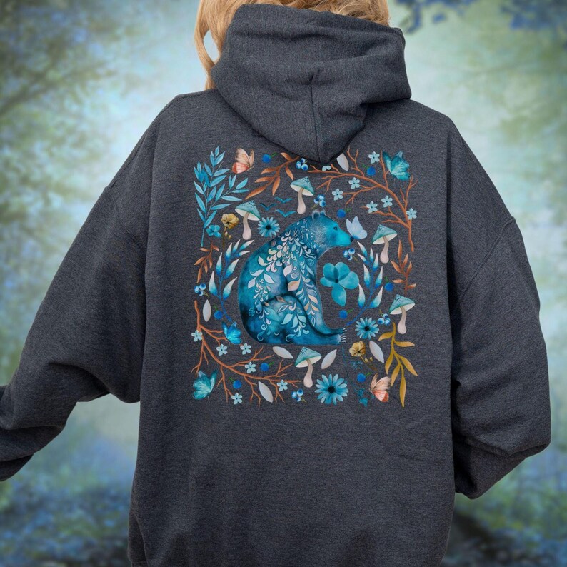 May include: Dark gray hoodie featuring a blue bear design surrounded by flowers, mushrooms, and butterflies. The bear is intricately detailed with floral patterns. The design is centered on the back of the hoodie, creating a nature-inspired aesthetic.