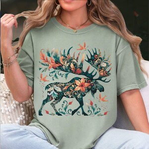 May include: Sage green t-shirt featuring a detailed illustration of a moose with ornate floral and leaf patterns in shades of orange, teal, and green. The moose has large antlers and is surrounded by stylized birds and foliage.