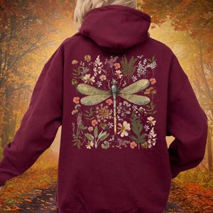 May include: Burgundy hoodie featuring a dragonfly design on the back. The dragonfly is surrounded by a floral arrangement with various flowers and greenery. The hoodie has a drawstring hood and a kangaroo pocket.
