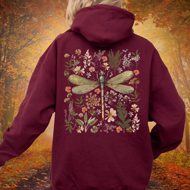 May include: Burgundy hoodie featuring a dragonfly design on the back. The dragonfly is surrounded by a floral arrangement with various flowers and greenery. The hoodie has a drawstring hood and a kangaroo pocket.