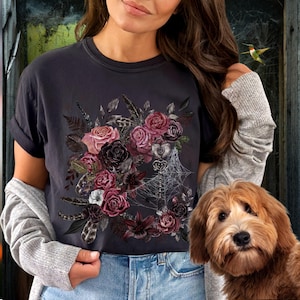 May include: A dark gray t-shirt with a floral design featuring dark and light pink roses, black feathers, and a spiderweb. The design is centered on the shirt.