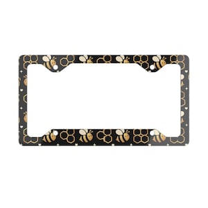 May include: Black license plate frame with a repeating pattern of gold honeycombs, bees, and small white hearts. The frame has a rectangular shape with cutouts for mounting.