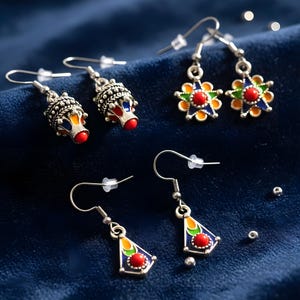 May include: A collection of silver-toned earrings with colorful enamel details. The earrings feature various designs, including a floral shape, a teardrop shape, and a detailed, ornate design. Each earring has a red, blue, yellow, and green color scheme.