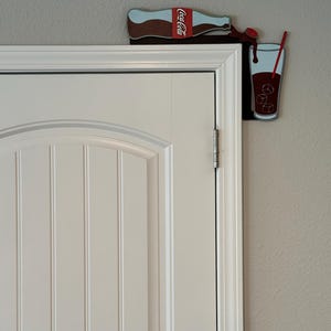 May include: A Coca-Cola bottle pouring into a glass with ice cubes and a red straw. The bottle and glass are attached to a white door frame.