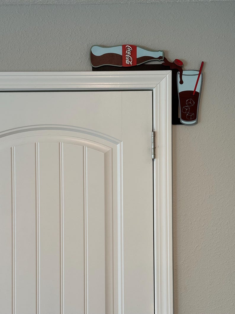 May include: A Coca-Cola bottle pouring into a glass with ice cubes and a red straw. The bottle and glass are attached to a white door frame.