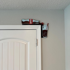 May include: A Coca-Cola bottle pouring into a glass with ice cubes and a red straw. The bottle and glass are attached to a wall, creating a fun and whimsical design.