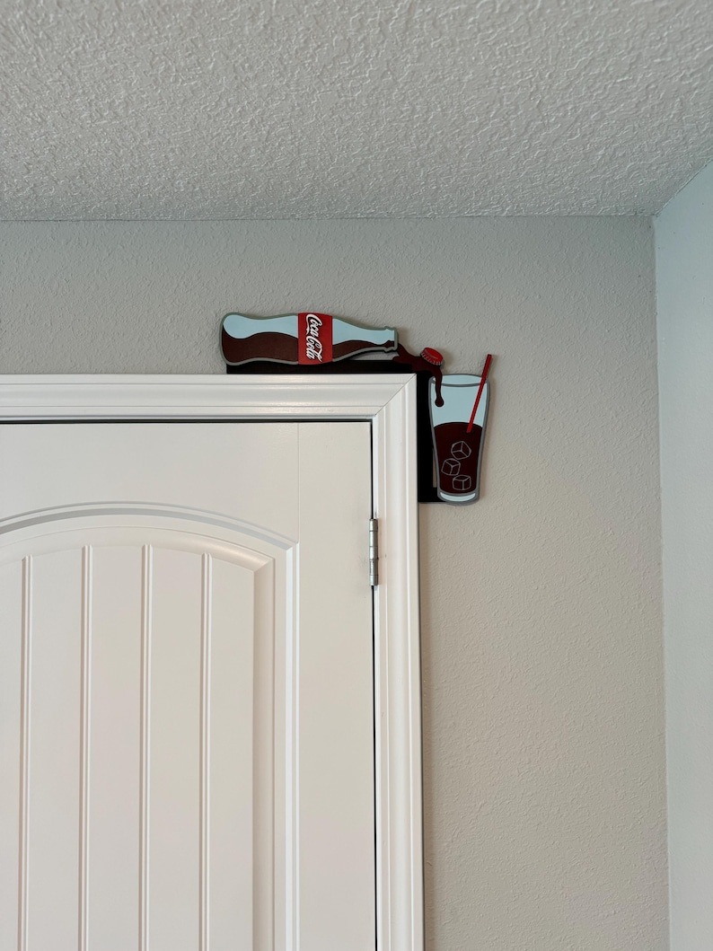 May include: A Coca-Cola bottle pouring into a glass with ice cubes and a red straw. The bottle and glass are attached to a wall, creating a fun and whimsical design.