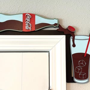 May include: A wooden cutout of a Coca-Cola bottle pouring into a glass with ice cubes and a red straw. The bottle is brown with a red Coca-Cola label. The glass is clear with a blue interior and a brown liquid. The cutout is mounted on a white door frame.