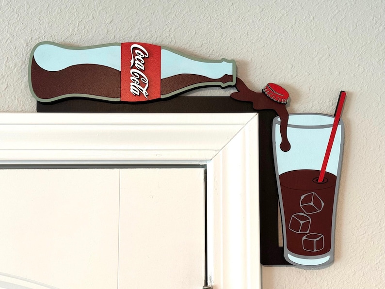 May include: A wooden cutout of a Coca-Cola bottle pouring into a glass with ice cubes and a red straw. The bottle is brown with a red Coca-Cola label. The glass is clear with a blue interior and a brown liquid. The cutout is mounted on a white door frame.