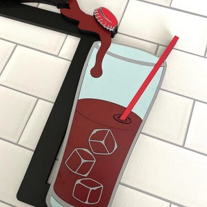 May include: A wooden cutout of a glass of cola with ice cubes and a red straw. The cola is being poured from a bottle with a red cap. The background is a white tiled wall.