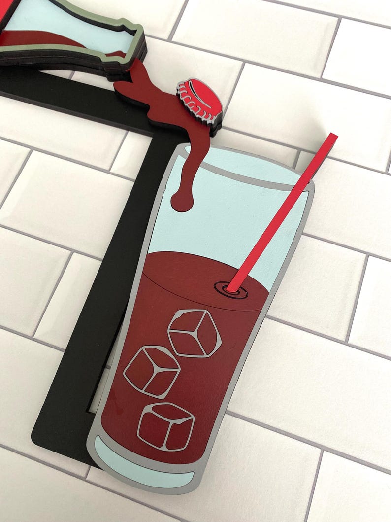 May include: A wooden cutout of a glass of cola with ice cubes and a red straw. The cola is being poured from a bottle with a red cap. The background is a white tiled wall.