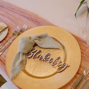 May include: A gold charger plate with a light green fabric napkin and a wooden name card that says "Blakely".