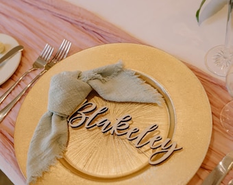 May include: A gold charger plate with a light green fabric napkin and a wooden name card that says "Blakely".