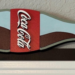 May include: A wooden wall decor shaped like a Coca-Cola bottle pouring out brown liquid. The bottle is painted blue and white with a red Coca-Cola label. The bottle is attached to a brown wooden base.