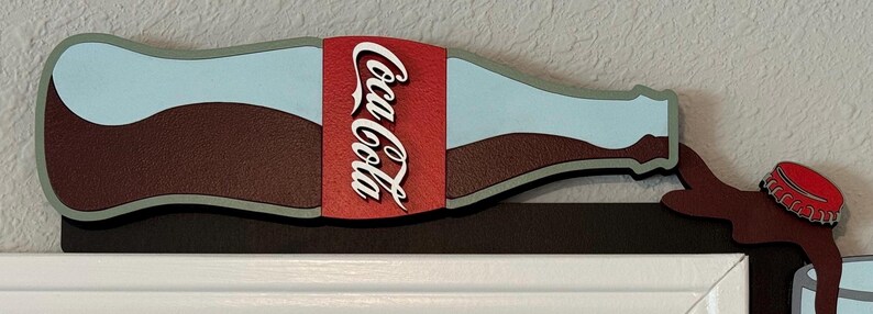 May include: A wooden wall decor shaped like a Coca-Cola bottle pouring out brown liquid. The bottle is painted blue and white with a red Coca-Cola label. The bottle is attached to a brown wooden base.