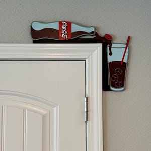 May include: A Coca-Cola bottle pouring into a glass with ice cubes and a red straw. The bottle and glass are attached to a white door frame.
