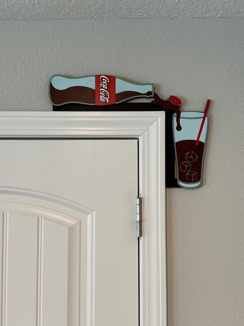 May include: A Coca-Cola bottle pouring into a glass with ice cubes and a red straw. The bottle and glass are attached to a white door frame.