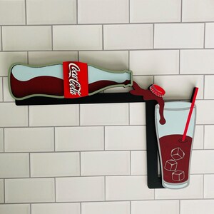 May include: A wooden wall decor featuring a red Coca-Cola bottle pouring into a glass with ice cubes and a red straw. The bottle has a white wave design and the glass is outlined in black.
