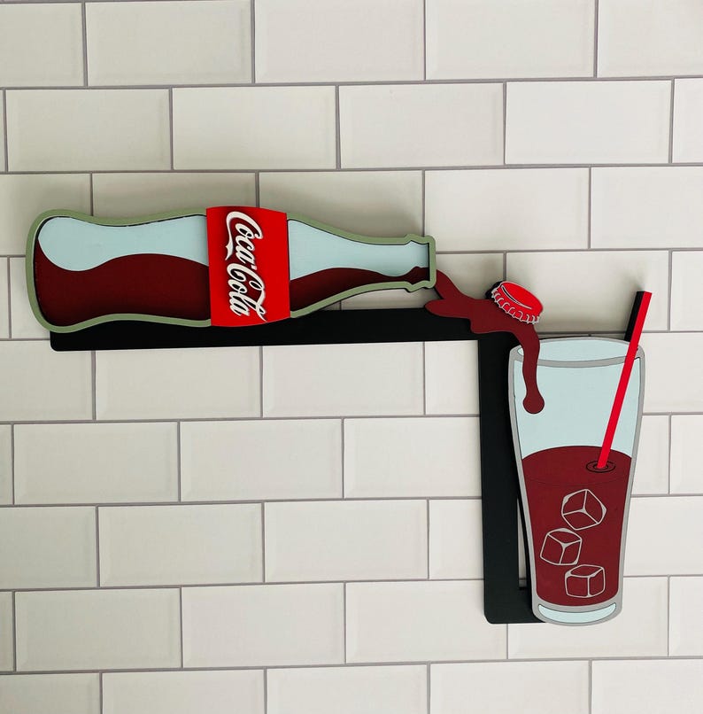 May include: A wooden wall decor featuring a red Coca-Cola bottle pouring into a glass with ice cubes and a red straw. The bottle has a white wave design and the glass is outlined in black.