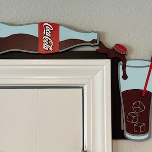 May include: A wooden wall decor featuring a Coca-Cola bottle pouring into a glass with ice cubes and a red straw. The bottle is brown with a red and white Coca-Cola label. The glass is blue with a brown liquid inside. The background is a white wall.