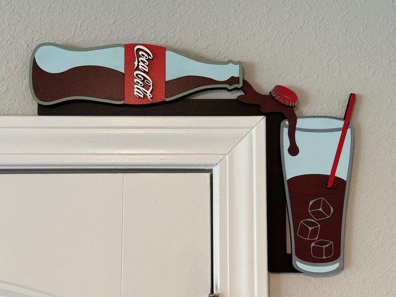 May include: A wooden wall decor featuring a Coca-Cola bottle pouring into a glass with ice cubes and a red straw. The bottle is brown with a red and white Coca-Cola label. The glass is blue with a brown liquid inside. The background is a white wall.