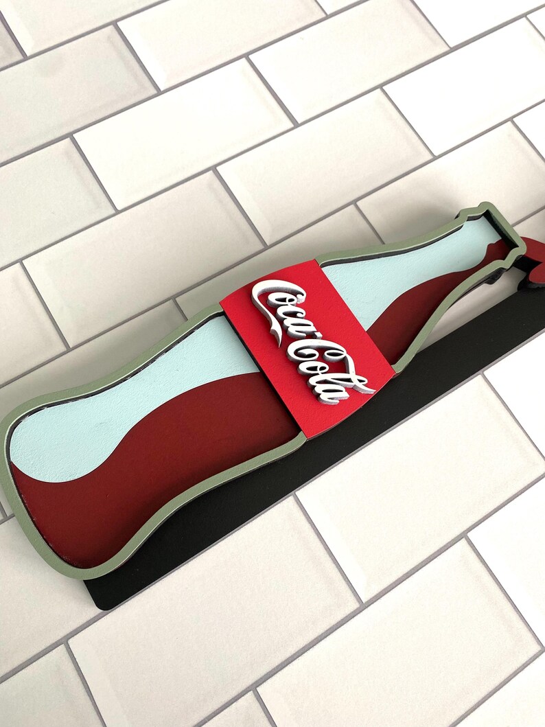 May include: A red, white, and green Coca-Cola bottle cutout with a black backing. The bottle is shaped like a classic Coca-Cola bottle and has the Coca-Cola logo on the label.