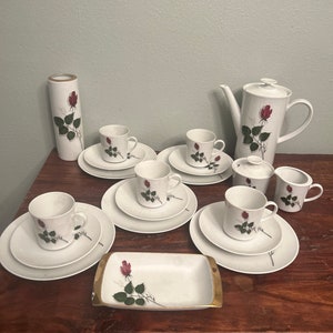 May include: A white tea set with a rose pattern. The set includes a teapot, creamer, sugar bowl, six cups, and six saucers. The set is on a wooden table.