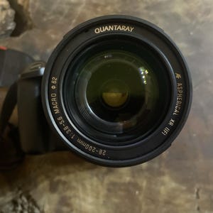 May include: A black Quantaray AF Aspherical XR IF lens with a 62mm macro and 28-200mm 1:3.8-5.6 specifications. The lens is attached to a black camera body with a Canon strap.